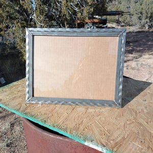 May include: A silver metal picture frame with a textured border. The frame is sitting on a wooden surface.