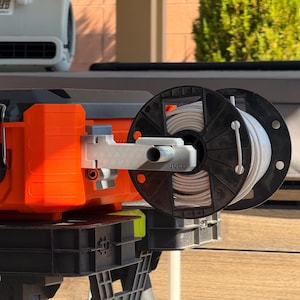 May include: An orange and black toolbox with a white spool of material attached. The spool is mounted on a gray arm, and the text "JUCO" is visible. A gray fan is in the background.