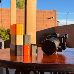 May include: A stack of four rectangular blocks in shades of orange, tan, and black, with the text "JUCOPRINT" and "The Bennetts" printed on the sides. A Sony camera with a large lens sits on a wooden table.
