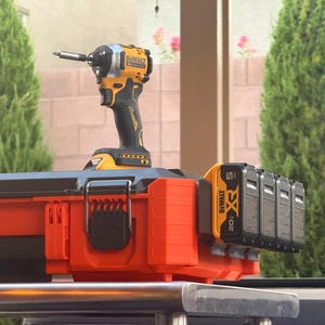 May include: An orange and black DeWalt impact driver with a silver bit is mounted on an orange and black toolbox. Five black 20V XR batteries are attached to the side of the toolbox. The tools are on a metal surface.