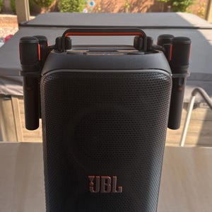 May include: Black portable speaker with a mesh front and the JBL logo in orange. It has a black handle with orange accents and includes four black microphones with orange details. The speaker is designed for karaoke and music.