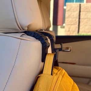 May include: Black car seat headrest hooks with a yellow jacket hanging from one. The hooks are made of a chain-like design and attach to the car seat. The jacket is a bright yellow color.