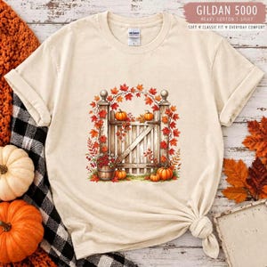 May include: A cream-coloured short-sleeve t-shirt featuring a graphic of a wooden gate adorned with orange pumpkins and autumn leaves. The shirt has a Gildan 5000 label at the neck. The design includes a small wooden bucket filled with berries.