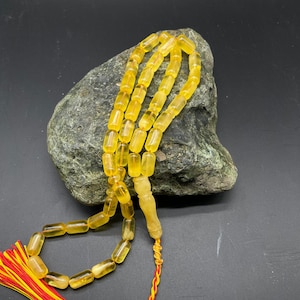 Op de afbeelding: Een gele amber gebedsketting met een kwast van rood en geel garen. De kralen zijn rechthoekig van vorm en zijn geregen aan een koord.