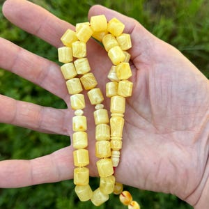 May include: A string of prayer beads featuring smooth, cylindrical, pale yellow amber-coloured beads. The beads are uniform in shape and size, connected by a thin cord, with a slightly larger, decorative carved bead positioned near the end.