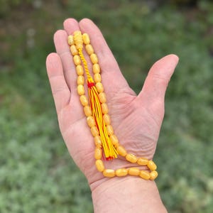 Puede incluir: Un rosario de cuentas de oración de color ámbar claro con una borla amarilla y roja. Las cuentas de forma ovalada están ensartadas con un cordón amarillo. La borla está hecha de hilos amarillos y rojos.