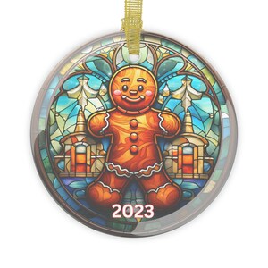 May include: Round Christmas ornament featuring a gingerbread man design with a stained glass background. The gingerbread man is orange with white icing details. The ornament has a gold ribbon for hanging and the year "2023" is printed on the bottom.