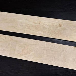 K&ouml;nnte beinhalten: Zwei rechteckige Holzbretter, beide hellbraun, mit nat&uuml;rlicher Holzmaserung. Die Bretter liegen flach auf einer schwarzen Oberfl&auml;che.