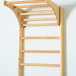 K&ouml;nnte beinhalten: Eine Wandmontage-Klimmzugstange aus Holz mit einer gebogenen Oberseite und mehreren horizontalen Stangen. Die Stange ist aus Naturholz gefertigt und hat eine hellbraune Oberfl&auml;che.