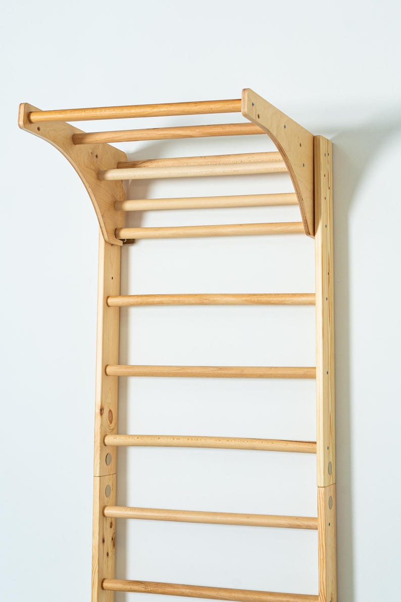 K&ouml;nnte beinhalten: Eine Wandmontage-Klimmzugstange aus Holz mit einer gebogenen Oberseite und mehreren horizontalen Stangen. Die Stange ist aus Naturholz gefertigt und hat eine hellbraune Oberfl&auml;che.