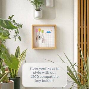 May include: A wooden key holder with a white baseboard and a LEGO compatible design. The key holder has a white frame and is mounted on a white wall. There are four keys hanging on the holder, and a blue cloud and colourful LEGO bricks are on the baseboard. The text "Store your keys in style with our LEGO compatible key holder!" is written in white text on a white background.
