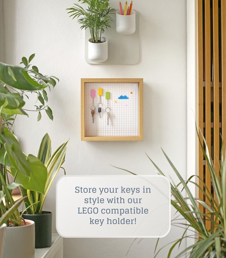 May include: A wooden key holder with a white baseboard and a LEGO compatible design. The key holder has a white frame and is mounted on a white wall. There are four keys hanging on the holder, and a blue cloud and colourful LEGO bricks are on the baseboard. The text "Store your keys in style with our LEGO compatible key holder!" is written in white text on a white background.