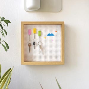 May include: A wooden frame with a white Lego-style brick wall inside. The wall has four colourful Lego bricks and three keys hanging on hooks. There are also small Lego bricks in various colours on the wall.