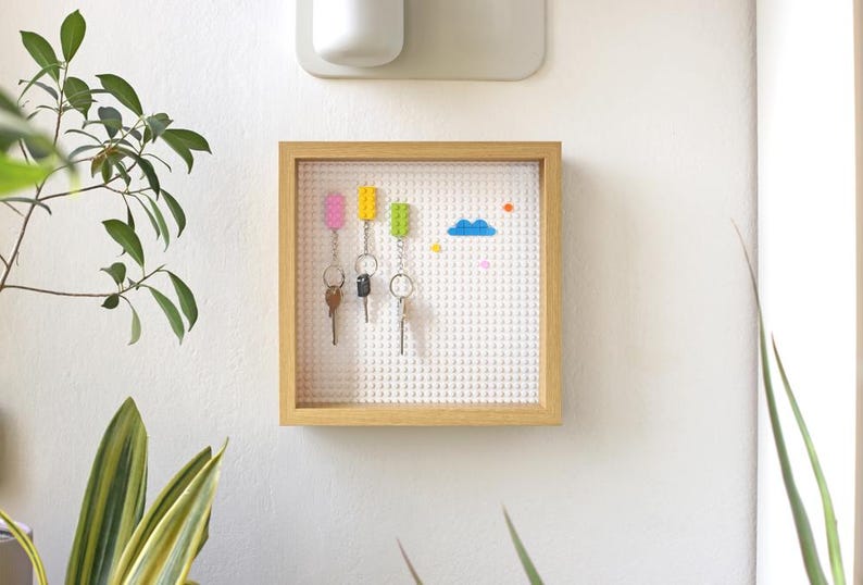 May include: A wooden frame with a white Lego-style brick wall inside. The wall has four colourful Lego bricks and three keys hanging on hooks. There are also small Lego bricks in various colours on the wall.