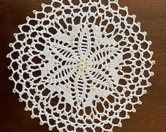 May include: White crocheted doily with a circular design and gold embellishments around the edge.