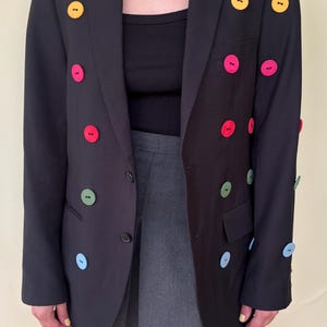 May include: A black blazer featuring large, round, multi-colored buttons in yellow, pink, red, green, and light blue arranged in rows on the front and sleeves. The blazer has a notched lapel, long sleeves, and two front flap pockets.