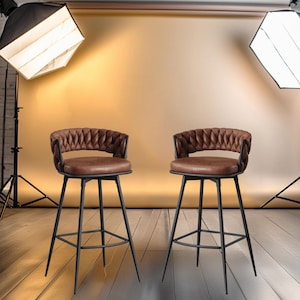 May include: Two brown leather bar stools with black metal frames and a woven back design. The stools are set against a backdrop of a brick wall and a white backdrop.
