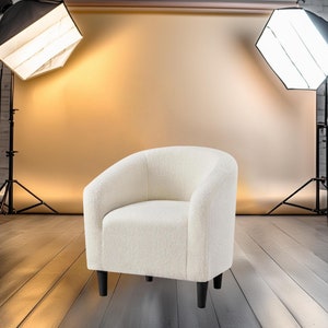 May include: A cream-colored, curved armchair with a textured fabric. The chair has a rounded back and black legs. The image is taken in a studio setting with lighting equipment and a backdrop. The text "Woody Floors" is in the top left corner.