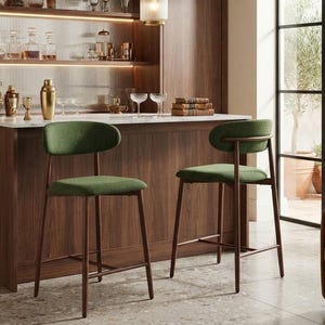 May include: Two bar stools with dark wood frames and forest green upholstered seats and curved backrests. The stools feature four slender legs connected by a footrest, positioned in front of a wooden home bar with a white countertop and shelving.