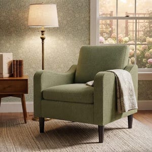 May include: An olive green upholstered armchair with dark brown wooden legs, featuring a textured fabric finish. A light-colored patterned throw blanket is draped over the right armrest, providing a soft contrast to the solid green seat and back.