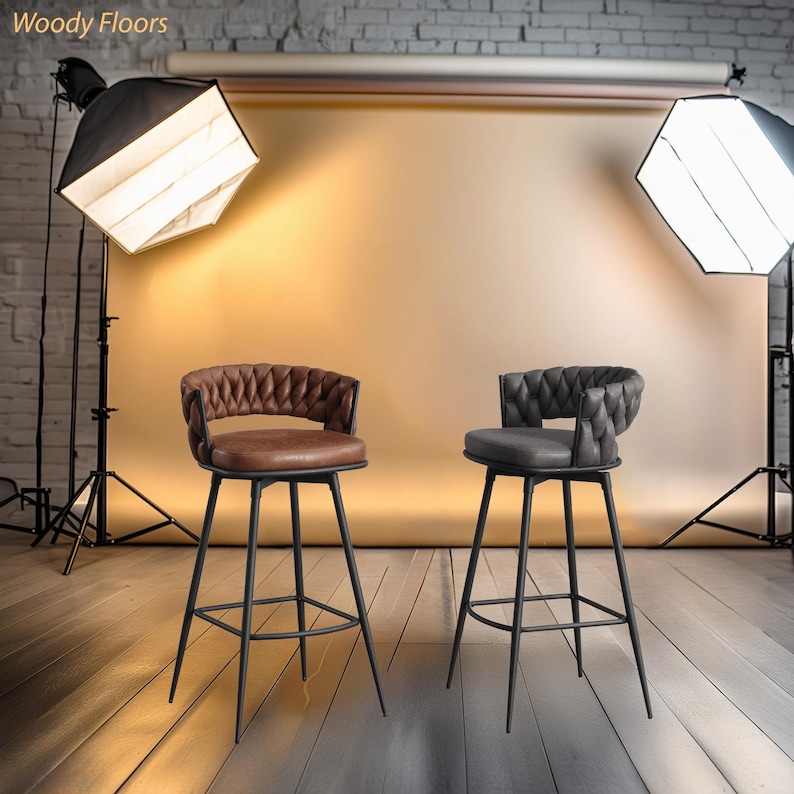 May include: Two black metal bar stools with brown and grey upholstered seats and a woven back design. The stools are set against a backdrop of a brick wall and a light brown backdrop.