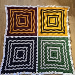 Può includere: Una coperta all'uncinetto con quattro quadrati di diversi colori: marrone, giallo, nero e verde. Ogni quadrato ha un bordo bianco e un quadrato più piccolo all'interno. La coperta ha un bordo bianco all'uncinetto.