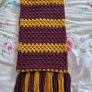 Può includere: Sciarpa all'uncinetto fatta a mano bordeaux e oro. La sciarpa presenta strisce alternate bordeaux e oro, con nappe abbinate alle estremità. Realizzata in filato morbido, è perfetta per l'inverno. Un accessorio caldo e colorato.