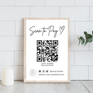 Può includere: Un cartello incorniciato con la scritta "Scan to Pay" in elegante corsivo, un codice QR e i nomi utente dei social media. Il cartello è in una cornice di legno chiaro, su uno sfondo di piastrelle bianche. Il cartello include anche il testo "APP NAME @BUSINESSNAME" e un indirizzo web.