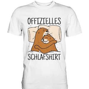 Könnte beinhalten: Ein weißes T-Shirt mit kurzen Ärmeln und einer Grafik von zwei braunen Ottern, die sich im Bett unter einer Decke mit Kissen umarmen. Der Text "OFFIZIELLES SCHLAFSHIRT" ist in schwarzen Großbuchstaben über und unter der Illustration gedruckt.