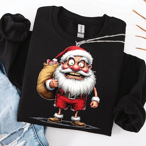 Könnte beinhalten: Schwarzes Sweatshirt mit einem Cartoon-Weihnachtsmann mit wütendem Gesichtsausdruck. Der Weihnachtsmann trägt einen roten Anzug und Hut und hält einen Geschenkesack. Das Sweatshirt wird auf einer weißen Oberfläche mit einer Jeans präsentiert.