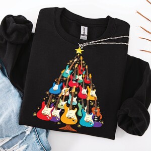 Könnte beinhalten: Ein schwarzes Langarm-Sweatshirt mit einem Grafikdruck eines Weihnachtsbaums, der aus verschiedenen bunten E-Gitarren besteht. Der Baum ist mit einem gelben Stern gekrönt und von kleinen goldenen Sternakzenten auf dem schwarzen Stoff umgeben.