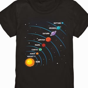 Könnte beinhalten: Schwarzes T-Shirt mit einer farbenfrohen Illustration des Sonnensystems. Das Design zeigt die Sonne und die Planeten: Merkur, Venus, Erde, Mars, Jupiter, Saturn, Uranus und Neptun, jeweils beschriftet. Die Planeten sind in ihren charakteristischen Farben dargestellt.