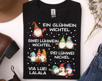 May include: A black short-sleeve t-shirt featuring a graphic of Christmas gnomes and the text: EIN GLÜHWEIN WICHTEL, SWEI LÜHWEIN WICHTEL, DEI LÜHWEI NICHEL, VIA LÜEI LALALA. The design includes white snowflakes scattered across the black fabric.