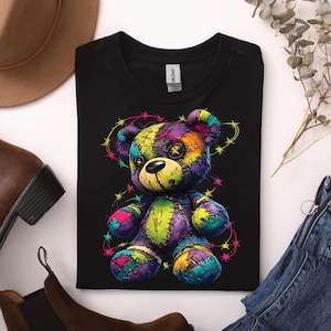 Könnte beinhalten: Schwarzes T-Shirt mit einem farbenfrohen Patchwork-Teddybär-Design. Der Bär hat ein gelbes Gesicht und Knopfaugen, umgeben von einem sternförmigen Muster. Das Design verwendet verschiedene Farben, darunter Lila, Grün und Rosa.