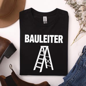 Könnte beinhalten: Ein schwarzes T-Shirt mit dem weißen Schriftzug "BAULEITER" über einer weißen grafischen Abbildung einer Trittleiter. Das T-Shirt hat einen Rundhalsausschnitt und eine Standardpassform, flach ausgelegt mit sichtbarem Markenetikett am Kragen.