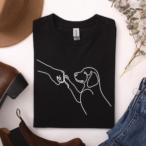 Könnte beinhalten: Schwarzes T-Shirt mit einer weißen Strichzeichnung eines Fistbumps mit einem Hund. Der Hund ist mit einer einzigen durchgehenden Linie dargestellt. Das Shirt besteht aus einem weichen Material.