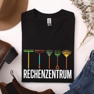 Könnte beinhalten: Ein schwarzes T-Shirt mit kurzen Ärmeln, das eine Grafik von fünf verschiedenen bunten Gartenrechen in einer Reihe über dem weißen Schriftzug RECHENZENTRUM zeigt. Das Shirt hat einen Rundhalsausschnitt und ein deutlich sichtbares Markenetikett.