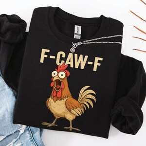 Könnte beinhalten: Ein schwarzes Sweatshirt mit Rundhalsausschnitt und einer Grafik eines braunen Huhns mit weit aufgerissenen Augen und offenem Schnabel. Über dem Huhn ist der Text F-CAW-F in einer hellbeigen Blockschrift gedruckt. Eine silberne Kette liegt am Ausschnitt.