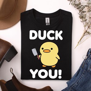 Könnte beinhalten: Ein schwarzes T-Shirt mit einer Grafik einer gelben Cartoon-Ente, die ein kleines silbernes Hackmesser hält. Der Text DUCK YOU! ist in großen weißen Großbuchstaben über und unter der Entenillustration auf der Vorderseite aufgedruckt.