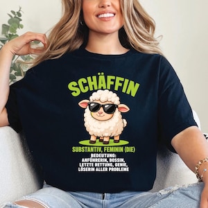 Könnte beinhalten: Ein schwarzes T-Shirt mit einer Grafik eines weißen Schafes, das eine schwarze Sonnenbrille trägt. Über dem Schaf steht der grüne Text "SCHÄFFIN". Darunter steht der grüne Text "SUBSTANTIV, FEMININ (DIE)" und der weiße Text "BEDEUTUNG: ANFÜHRERIN, BOSSIN, LETZTE RETTUNG, GENIE, LÖSERIN ALLER PROBLEME".