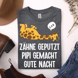 Könnte beinhalten: Ein dunkelgrau meliertes T-Shirt mit Rundhalsausschnitt und einer Grafik einer gelb-braunen Giraffe, die auf dem Rücken liegt, mit einer weißen Sprechblase und dem Text zzz. Darunter steht in weißer Schrift ZÄHNE GEPUTZT PIPI GEMACHT GUTE NACHT.