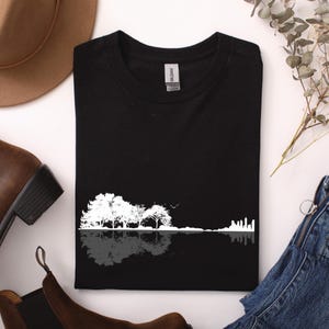 Könnte beinhalten: Schwarzes T-Shirt mit einem weißen Grafikdesign von Bäumen und einer Skyline, die sich im Wasser spiegelt. Das Design ist eine Silhouette. Das T-Shirt ist sauber gefaltet.