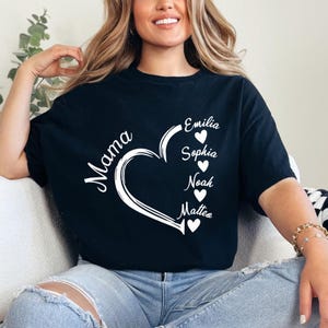 Op de afbeelding: Zwart t-shirt met een wit hartontwerp en het woord "Mama" in cursief. Het hart bevat de namen Emilia, Sophia, Noah en Matteo. Het t-shirt wordt door een persoon gedragen.