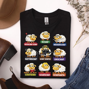 Könnte beinhalten: Schwarzes T-Shirt mit Cartoon-Schafen und deutschen Sätzen. Jedes Schaf hat ein einzigartiges Design und Etikett, darunter "Gestochen Schaf" und "Messerscharf". Das T-Shirt ist gefaltet.