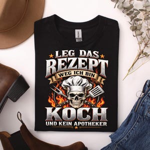 Könnte beinhalten: Ein schwarzes T-Shirt mit einem Grafikdruck eines Totenkopfes mit Kochmütze, platziert vor gekreuzten Kochutensilien und Flammen. Der Text lautet: LEG DAS REZEPT WEG ICH BIN KOCH UND KEIN APOTHEKER, in fetter, metallisch wirkender Schrift.