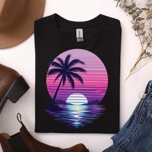 Könnte beinhalten: Schwarzes T-Shirt mit einem Sonnenuntergang-Design. Die Grafik zeigt eine Palmensilhouette, eine untergehende Sonne und Wasser in Lila-, Rosa- und Blautönen. Das Shirt ist gefaltet, mit einem braunen Hut und Stiefeln im Hintergrund.