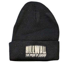 Może przedstawiać: Czarna dzianinowa czapka z wywiniętym brzegiem. Z przodu czapki znajduje się prostokątna naszywka z napisem "MILLWALL" nad "THE PRIDE OF LONDON" w białym kolorze.