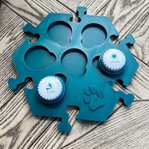 May include: Teal puzzle-shaped dog treat dispenser with six circular openings. Two white and blue lids are visible, one with a paw print and the word "Play", the other with a sun icon and the word "Outside". A paw print is also on the dispenser.