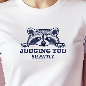 May include: White t-shirt with a black and white illustration of a raccoon peeking over the top and the text "JUDGING YOU SILENTLY."