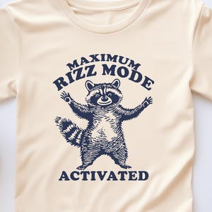 May include: A cream colored t-shirt with a raccoon graphic and the text "Maximum Rizz Mode Activated" in navy blue.